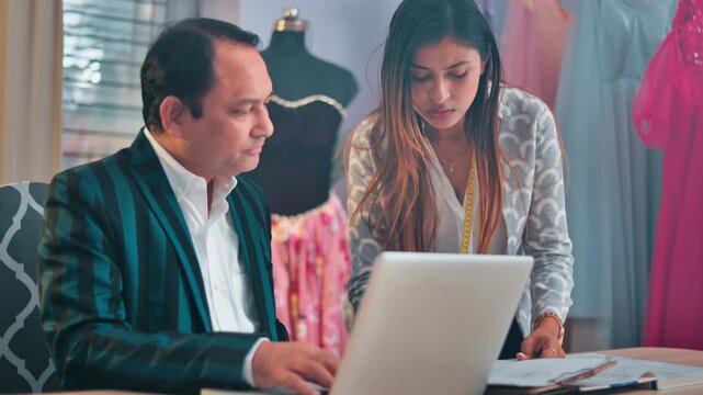 Indian female fashion designer discussing clothing ideas with a male designer while working on a garment project using a laptop in a fashion studio.