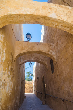 Overhead arches and vintage lanterns on a narrow street with the fortress wall in the background in the Cittadella, Victoria - Gozo MALTA