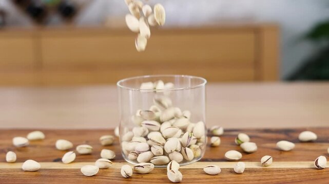 Slow motion footage of pistachio nuts falling into glass container on wooden surface.