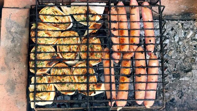 Close-up of grilled pork sausages and sliced ​​chicken rolls cooking together on a metal grate over hot coals outdoor, smoky flavored cooking during a summer picnic.