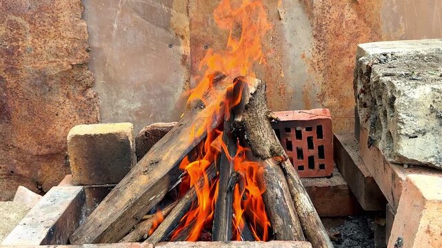Wood logs burning with bright orange flames inside a rustic stone and brick fire pit outdoors, close up shot of a crackling bonfire used for cooking or heating in a garden.