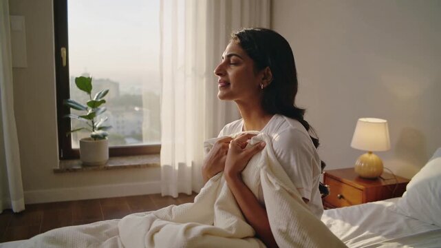Morning light drawing Indian woman wrapping cream blanket on bed, wearing white tee, hoop earrings