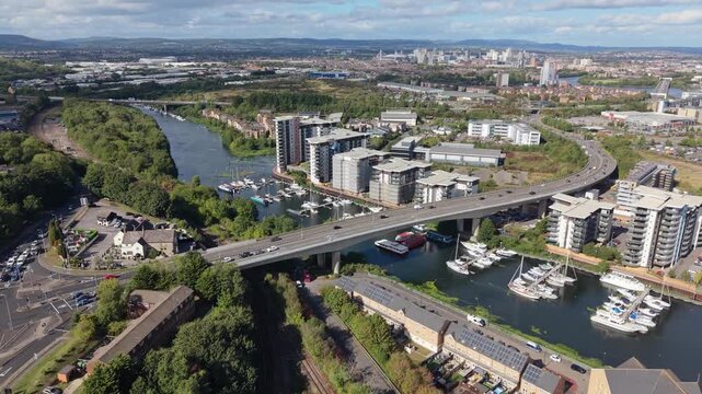 Wide aerial view of a waterfront district in England with a busy road bridge, marina, moored boats, modern apartment buildings and an extensive urban landscape in the distance.
