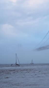 View of 25 de Abril Bridge famous tourist landmark of Lisbon connecting Lisboa and Almada in heavy fog mist wtih yacht boats passing under. Lisbon, Portugal