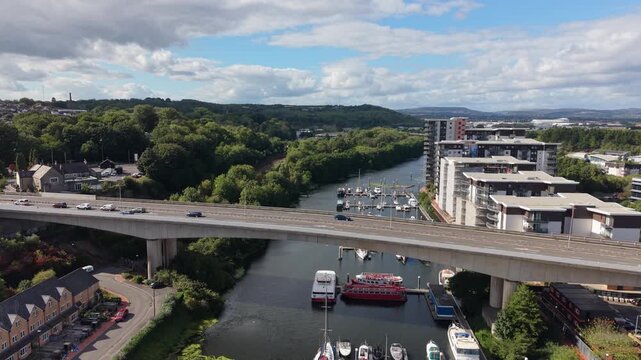 Drone flies toward a busy elevated road bridge over a waterway in England, with moving traffic, moored boats, marina, modern apartments and green surroundings.