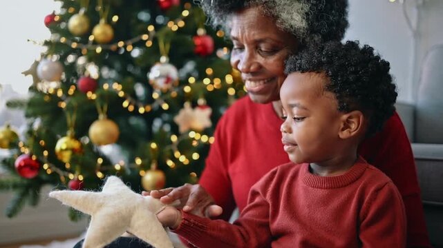 Presenting senior guiding child holding plush star at home by Christmas tree to hang, red sweaters