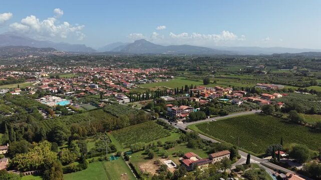 Town of Lazise spreads across green valley near Lake Garda beneath layered mountains, terracotta rooftops mix with vineyard rows, olive groves, scattered hillside villas. Aerial drone dolly forward