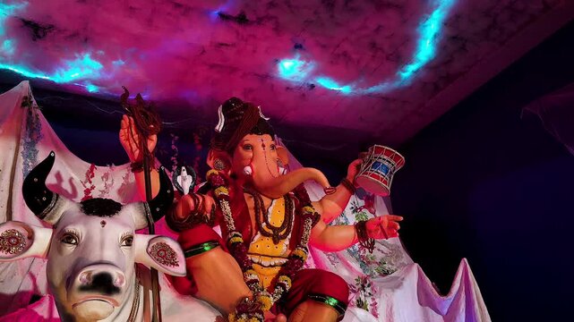 Ganesh idol dressed as Lord Shiva sits on Nandi bull, tilt shot reveals colorful lightning effect in decorative cloud ceiling during Ganesh Chaturthi pandal India.
