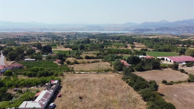 High altitude panoramic drone shot capturing green crop fields, farmhouses, and Polyfytos Lake on the horizon in Kozani region.