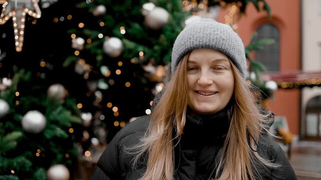 A cheerful blonde girl in a winter hat closing her eyes and smiling joyfully in front of a decorated holiday tree with warm glowing lights.