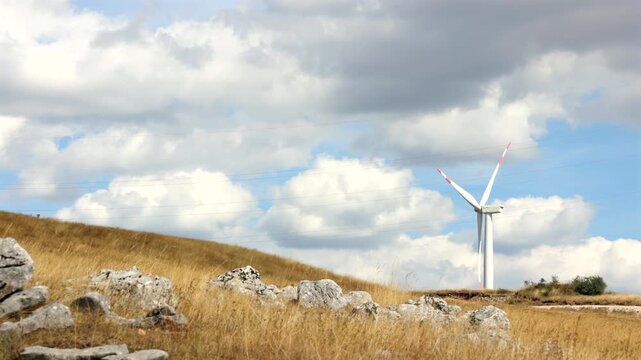 Wind Turbine on Golden Hills Under Cloudy Sky. Wind turbine standing on a dry grassy hillside with rocks and large clouds in the background. Renewable energy landscape in a rural mountain area.