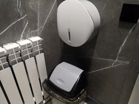 Round white paper towel dispenser is mounted on a dark marble-patterned tile wall above a small plastic trash bin in a modern, clean bathroom interior next to a white heating radiator.