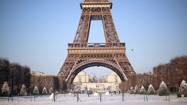 Front view of the Eiffel Tower rising above a snow-covered park in Paris on a sunny winter day. Blue sky, snowy grounds, and trimmed trees create a postcard-perfect scene.