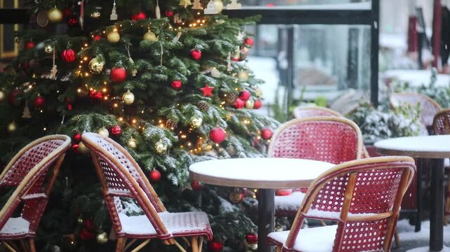 Cozy Parisian cafe terrace with red rattan chairs and round tables covered in fresh snow. A beautifully decorated Christmas tree with lights and ornaments stands in background as snow falls heavily.