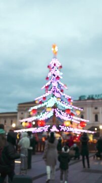 Beautiful tall outdoor Christmas tree adorned with colorful ornaments and bright lights standing at a festive holiday market on a winter afternoon.