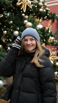 A cheerful young blonde woman in a grey knitted beanie hat and black winter jacket waving her hand and smiling on a festive decorated city street.