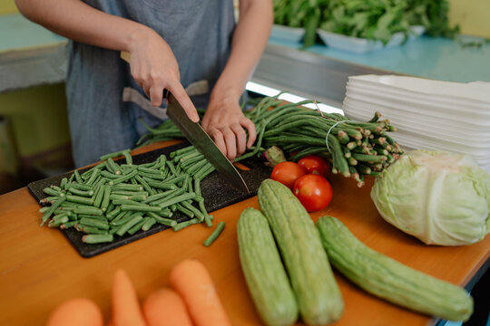 Hands prepare fresh vegetables on countertop, cutting green beans with knife beside tomatoes, cucumbers, cabbage, carrots and stacked trays in bright kitchen setting for healthy cooking.