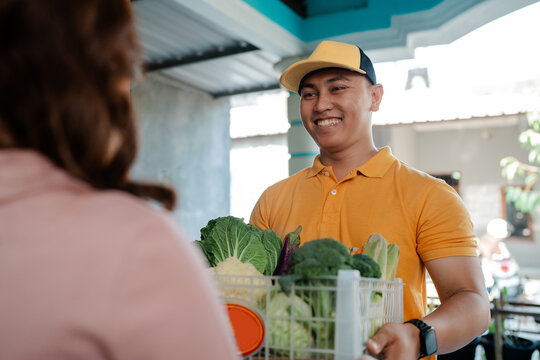 Smiling man in orange shirt and cap presents basket filled with fresh vegetables to customer in bright indoor market space, showing friendly service, local food shopping, and healthy produce exchange.