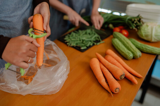 Close view of hands peeling carrot into plastic bag placed near green beans, cucumbers, tomatoes, and cabbage on wooden kitchen table during meal prep.