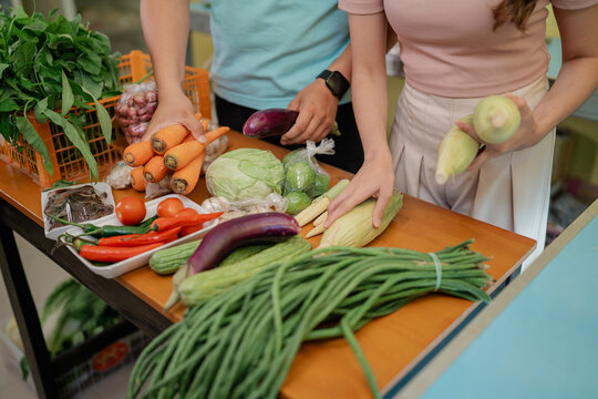 Shoppers reach over wooden market table to pick carrots, eggplants, cabbage, corn, chilies, tomatoes, and green beans, supporting healthy meal preparation and local food buying.