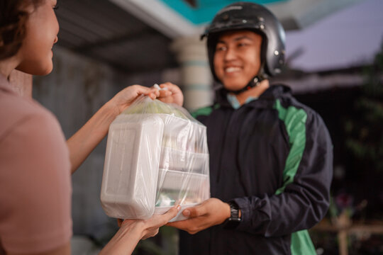 Woman hands boxed meal in clear plastic carry bag to smiling helmeted courier outdoors, showing local food delivery handoff, customer service, convenience, and fast urban service in soft evening light