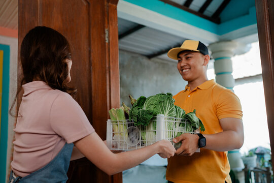 Smiling young man in orange cap and shirt offers leafy greens crate to woman at home doorway, showing friendly neighborhood delivery of fresh healthy produce for everyday sharing together.