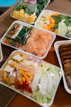 Overhead view of assorted hot pot ingredient trays with sliced meat, tofu, vegetables, mushrooms, corn, cabbage, carrots, broccoli, cauliflower, greens, and seasonings ready for shared cooking at home