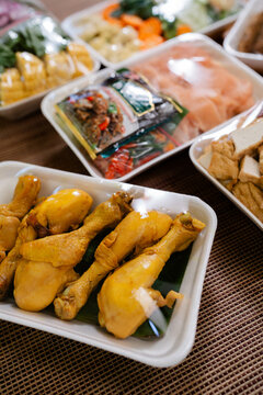 Cooked chicken drumsticks sit in foam trays beside assorted packaged ingredients and fresh produce on tabletop, creating warm close up view of ready to serve food for market, catering or buffet use.