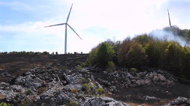 Wind turbines standing above a scorched rocky landscape with smoke rising near the forest after a wildfire. Renewable energy infrastructure in a fire-damaged natural area.
