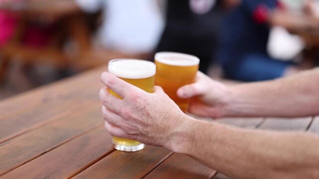 Hands placing two glasses of cold craft beer on wooden table in outdoor garden seating area during daytime.