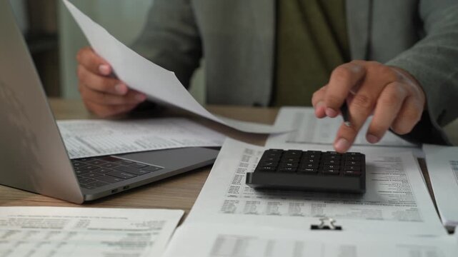 Businessman typing on laptop beside printed spreadsheets and financial reports, showing accounting spreadsheet analysis, data review and business reporting.