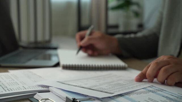 Businessman taking notes beside printed spreadsheets and laptop, showing accounting document review, financial reporting and office paperwork analysis.