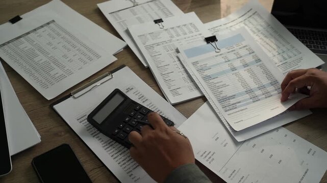 Overhead view of hands reviewing printed spreadsheets beside calculator, showing accounting report analysis, financial document checking and business paperwork.