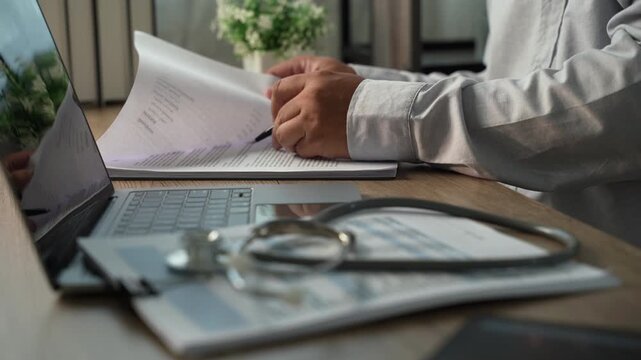 Hands reviewing medical paperwork with pen beside laptop and stethoscope, showing medical insurance document review, healthcare expense analysis and records checking.