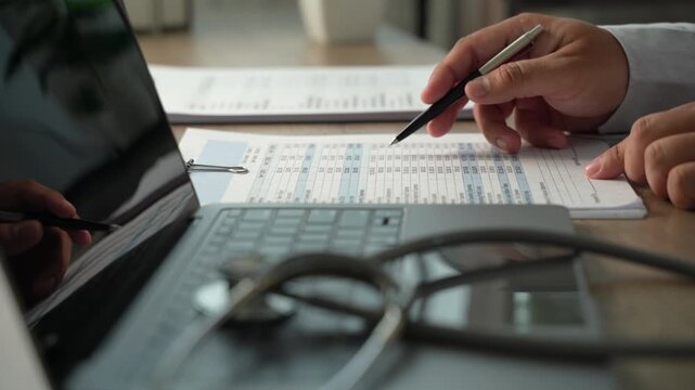 Hands reviewing medical paperwork with pen beside laptop and stethoscope, showing medical insurance document review, healthcare expense analysis and records checking.