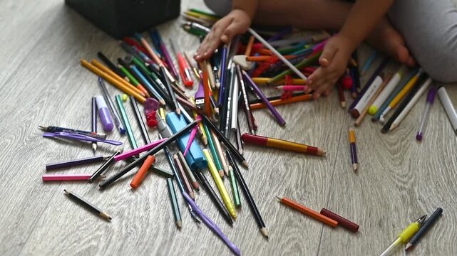 Pencil scattered across wooden floor around child hands, colorful pencil pile from open box creating playful art mess. drawing activity, creativity, education concept