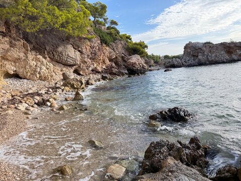 Calm waves washing onto a pebble and rock beach, framed by a steep cliff face with green pine trees, creating a peaceful natural coastal scene on a sunny day in calvia, spain