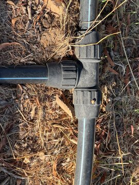 Black polyethylene irrigation pipe with t connector laid on dry soil and dead leaves, close up showing water distribution hardware for drought resilient agricultural water management, copy space