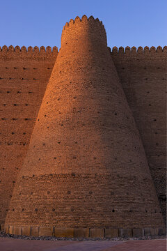 Conical brick tower of the Ark Fortress in Bukhara bathed in golden sunset light against a clear blue sky. Ancient Central Asian fortification wall with crenellated battlements, ideal for travel.