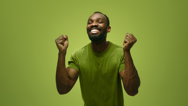 Victorious man raises clenched fists in green studio with triumphant excitement. Achievement, winning moment, motivational content concept.