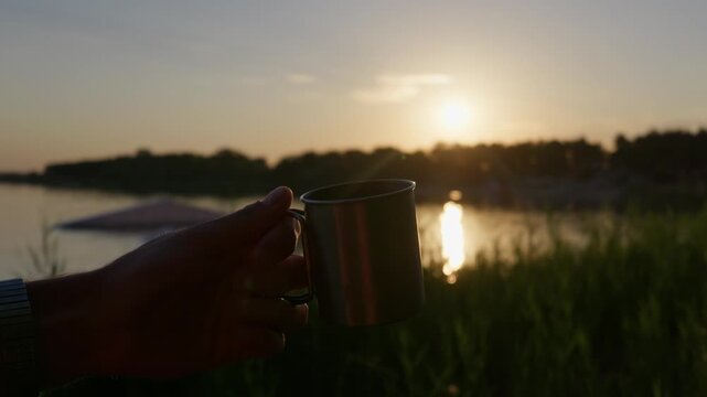 Hand holds a durable metal mug against a glowing sunset by the river. Warm evening light outlines the steel container beside lakeside vegetation during a calm twilight in nature.