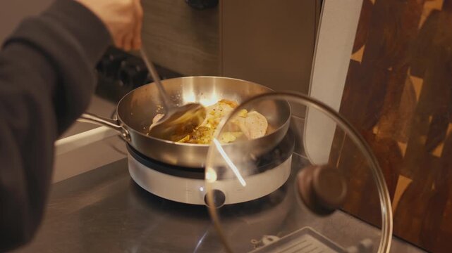 Close-up of woman hands turning sizzling chicken wings with a metal spatula in a stainless steel frying pan. Cooking poultry with ginger and garlic on a portable induction cooktop in a home kitchen.