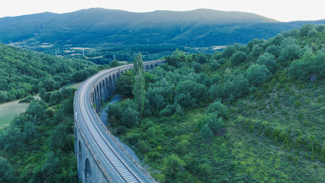 Elevated train tracks on a stone viaduct spanning a lush green valley with surrounding wooded hills and distant mountains