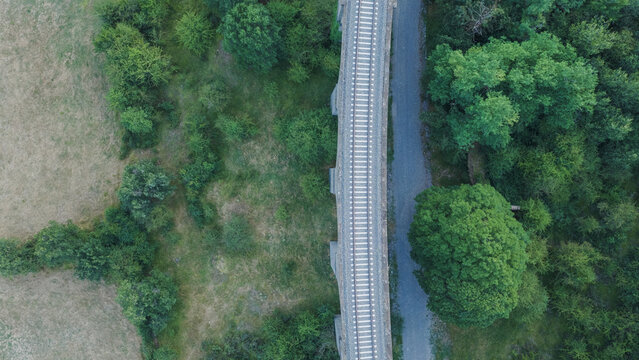 Elevated railway tracks crossing a lush green landscape and dry field, with a narrow path running alongside. Transportation infrastructure in nature