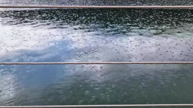 Raindrops falling on a serene lake creating concentric water ripples viewed through boat railing cables. Concept of rainy weather, stormy atmosphere, and climate change.