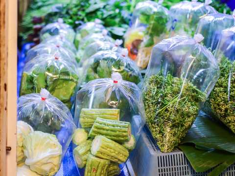Fresh raw vegetables in tied clear plastic bags at a Thai fresh market, including Malabar spinach shoots, bitter gourd and small cabbage heads, portioned on banana leaf for daily home cooking.