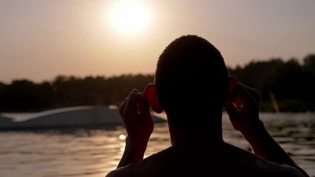 Man raises fists celebrating while standing in calm lake waters facing the bright setting sun. Warm backlighting creates glowing rim highlights and tranquil scenic solitude.