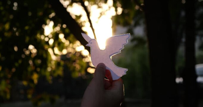Hand holds white dove cutout in forest sunlight. Golden rays shine through paper bird with lens flare and peaceful hope symbolism. Scene represents freedom, faith, healing, and spiritual peace.