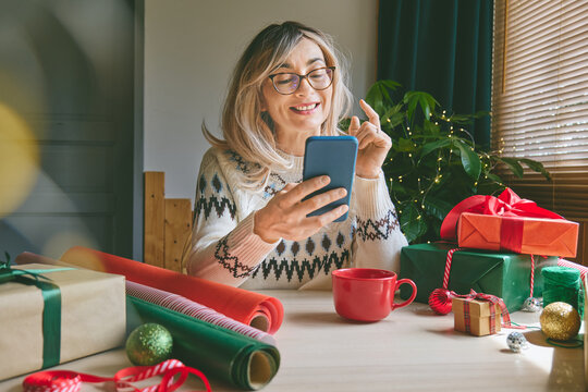 Woman using a smartphone while preparing Christmas gifts at home. She checks her phone during holiday preparations, suggesting online shopping, e-commerce, digital communication and seasonal gifting.