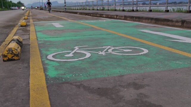 Static low angle shot of cyclists riding on a green bicycle lane along the Mekong River at sunset, Thailand.